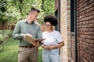 smiling real estate agent showing documents to african american woman outside of home.
